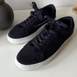 Marinblåa sneakers från Les Deux - Snygga marinblåa sneakers i mocka från Les Deux med vita sulor och snörning. Diskreta broderade detaljer i rött och blått på hälen ger en stilren touch. Perfekta för en avslappnad look. Storlek 42. De är i mycket bra skick. 