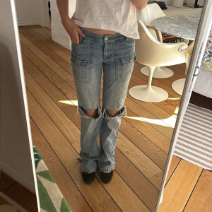 Blå jeans med slitningar - Säljer ett par blå jeans med slitningar och hål på knäna. Byxorna har en normal passform med flera fickor och en knappgylf. Perfekta för en avslappnad stil. Slitna längst ner 