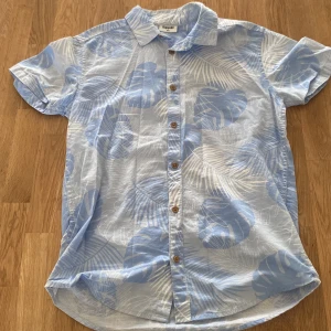 Blå kortärmad skjorta från Kappahl - Snygg blå skjorta från Kappahl med tropiskt bladmönster i vitt. Skjortan har korta ärmar och knappar framtill. Perfekt för en avslappnad sommarstil. 🌴