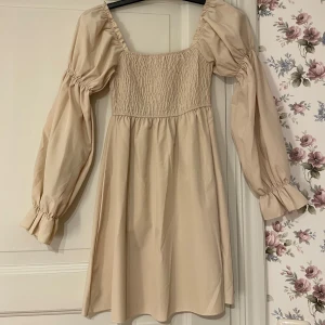 Beige klänning med puffärmar - Säljer en söt beige klänning med puffiga långa ärmar och smockdetaljer vid bysten. Perfekt för en stilren och romantisk look. Använd en gång.
