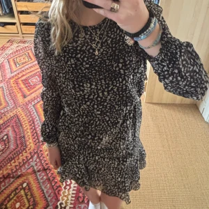 Svart och vit mönstrad klänning - Säljer en snygg svart och vit mönstrad klänning med volangdetaljer. Klänningen har långa ärmar och en kort längd. Den är använd få gånger och är i mycket bra skick.