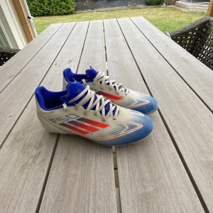 Adidas fotbollsskor i vitt, blått och rött - Snygga Adidas fotbollsskor i vitt med blå och röda detaljer. Skorna har snörning och en sportig design som passar perfekt för fotbollsplanen.