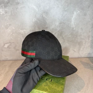 Svart Gucci keps - Hej! Säljer denna knappt använda svarta keps från Gucci med ett diskret mönster och klassiska gröna och röda ränder på sidan. Kepsen har ett justerbart spänne bak för en perfekt passform. Levereras med GG-lådan.                         Storlek- M 58Cm.                                                       För mer information kontakta oss gärna privat.