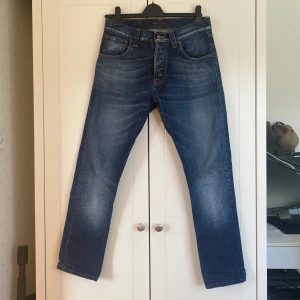 Slim jeans från Nudie Jeans - Säljer ett par snygga blå jeans från Nudie Jeans med klassisk femficksdesign och orange sömmar. Jeansen har en straight/slim passform och är tillverkade i slitstarkt denim. Perfekta för en avslappnad stil. Det står att de är W29/L32 men de är inlämnade hos skräddare så de är W29/L30. Dom är även insydda på benen för att bli mer slimmade.