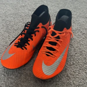 Orange och svarta fotbollsskor från Nike - Snygga fotbollsskor från Nike i en iögonfallande orange färg med svarta och silverdetaljer. Skorna har snörning och en rund tå, perfekt för fotbollsplanen. De har en slitstark sula med dobbar för optimalt grepp.