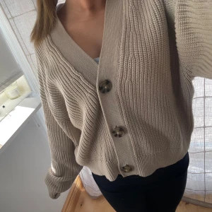 Beige stickad kofta från H&M - Säljer en mysig beige stickad kofta från H&M med stora knappar framtill. Koftan har en avslappnad passform och långa ärmar, perfekt för kyliga dagar. Den är både stilren och bekväm, ett måste i garderoben!