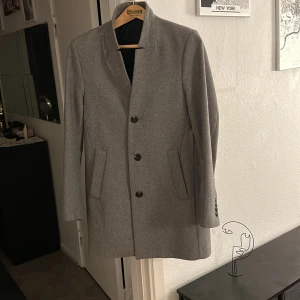 Grå kappa med knappar - Snygg grå kappa med stilrena knappar framtill. Tillverkad i en ullblandning för en elegant look. Perfekt för kyligare dagar med sin långa design och praktiska fickor.