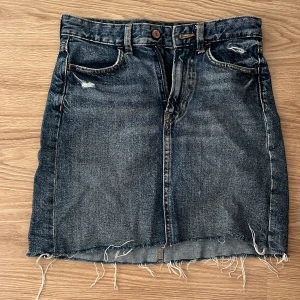 Jeanskjol från &Denim - Säljer en cool jeanskjol från &Denim i storlek 34. Kjolen har en rå kant nertill och slitna detaljer för en trendig look. Den är mörkblå med klassiska femficksdesign och stängs med dragkedja och knapp. Perfekt för en avslappnad stil!