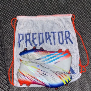 Adidas Predator Edge+ Fotbollsskor - Snygga Adidas Predator Edge+ fotbollsskor i en färgglad design med inslag av vitt, blått, rött och gult. Skorna har en strumpliknande passform utan snörning för en tight och bekväm känsla på planen. Perfekta för att dominera spelet med stil. Skorna e aldrig andvända och är 100% äkta. Jag säljer dom för dom inte passa och det gick inte att lämna tillbaka dom. 