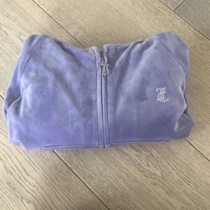 Lila jacka från Juicy Couture - Säljer en lila jacka från Juicy Couture med dragkedja och glittrande detaljer. Perfekt för en avslappnad och stilren look. Jackan har en broderad logga på framsidan och texten 'Juicy' i strass på baksidan. Kort i modellen! Liten i storleken. 