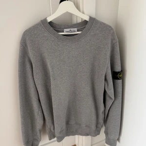 Grå sweatshirt från Stone Island - Säljer en stilren grå sweatshirt från Stone Island. Tröjan har en klassisk rund halsringning och ett ikoniskt märke på ärmen. Perfekt för en avslappnad och trendig look.