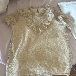 Beige spetsblus från Name It - Säljer en söt beige spetsblus från Name It. Blusen har korta ärmar och fina volangdetaljer vid halsen. Perfekt för en elegant look. Passar storlek 11-12 år.