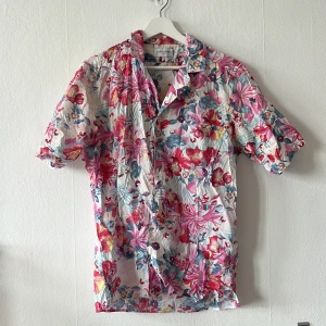 Blommig kortärmad skjorta från John Henric - Säljer en färgglad blommig skjorta från John Henric. Skjortan har korta ärmar och ett livfullt mönster i rosa, rött och blått på en vit bakgrund. Perfekt för sommarens alla tillfällen! Storlek M