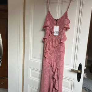 Rosa långklänning med volang från ZARA - Säljer en romantisk rosa långklänning från ZARA med tunna axelband och vackra volangdetaljer framtill. Klänningen har ett subtilt blommönster och är tillverkad i ett lätt, flowy material som ger en feminin siluett.