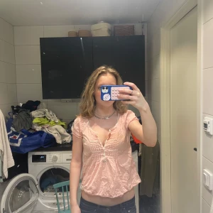 Ljusrosa blus med spetsdetaljer - Söt ljusrosa blus med korta ärmar och dekorativa spetsdetaljer längs halsringningen och knäppningen framtill. Blusen har små knappar och ett lätt, luftigt tyg som passar perfekt till varmare dagar.
