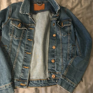 Blå jeansjacka från Lager 157 - Klassisk blå jeansjacka från Lager 157 med knappar framtill och på bröstfickorna. Jackan har långa ärmar, kontrastsömmar och två sidofickor. Perfekt för lager-på-lager och enkel att styla till olika outfits.