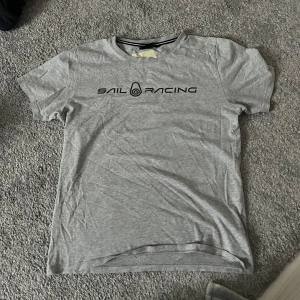 Grå t-shirt från Sail Racing - Säljer en grå t-shirt från Sail Racing med svart logga tryckt på bröstet. T-shirten har rund hals och korta ärmar. Perfekt för en avslappnad stil.