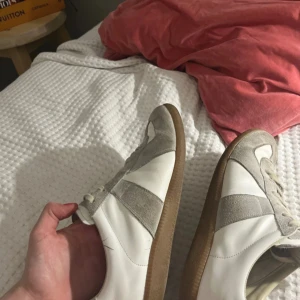 Maison Margiela Gats  - Säljer ett par klassiska Maison Margiela sneakers med vit läderbas och grå mockadetaljer. Skorna har bruna gummisulor och snörning framtill. Perfekta för dig som gillar stilrena och tidlösa sneakers. Kan tänka mig att byta. Pris ej hugget i sten 🪨 