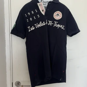 Marinblå pikétröja Les Voiles de St-Tropez - Säljer en marinblå pikétröja från Les Voiles de St-Tropez med broderade detaljer, tryck och patchar på både fram- och baksida. Tröjan har korta ärmar, krage och knappar framtill. Perfekt för en sportig och stilren look.