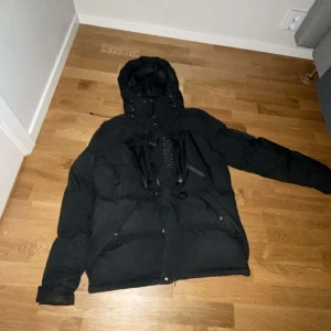 Svart pufferjacka från Hoodrich - Säljer en svart pufferjacka från Hoodrich i storlek S. Jackan har huva, dragkedja och knappar framtill samt två stora fickor. Perfekt för kyliga dagar och har en modern streetwear-look. Jag köpte den här jacken från JD sports för 1200. Priset kan diskuteras 