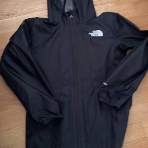 Svart vindjacka från The North Face - Svart vindjacka med huva från The North Face. Jackan har dragkedja framtill, elastiska muddar och meshfoder på insidan. Perfekt för blåsiga dagar och enkel att ta med sig. Klassisk logga på bröstet.
