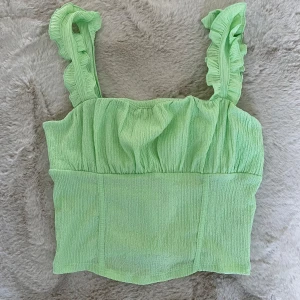 Grön croppad linnetopp med volang L oanvänd  - Säljer en ljusgrön croppad linnetopp från Gina Tricot. Toppen har breda axelband med volangdetaljer och är tillverkad i ett strukturerat, lätt material. Perfekt för varma dagar och enkel att styla med jeans eller kjol.
