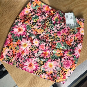 Blommig minikjol från H&M - Färgglad minikjol från H&M med stort blommönster i rosa, orange, grönt och gult. Kjolen har en rak passform och dragkedja bak. Tillverkad av 99% Lenzing Ecovero viskos och fodrad med 100% återvunnen polyester.