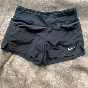 Svarta träningsshorts från Nike - Svarta träningsshorts från Nike med två framfickor och vit logga på vänster ben. Shortsen har en normal passform och är tillverkade i ett lätt syntetmaterial som passar perfekt för träning.