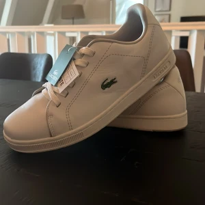 Vita sneakers från Lacoste - Säljer ett par nya fräscha vita sneakers från Lacoste med klassisk krokodillogga på sidan. Skorna har snörning, rund tå och är tillverkade i skinn. Perfekta för en stilren look och passar till många outfits.