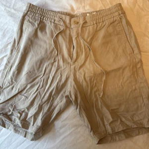 Beige shorts från NN07 med snörning - Snygga beige shorts från NN07 med elastisk midja och snörning. Modellen har fickor fram och bak samt stängs med knapp och dragkedja. Perfekta för varma dagar och har en avslappnad passform.