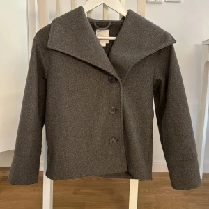Grå kappa från Gina Tricot - Säljer en stilren grå kappa från Gina Tricot. Den har en snygg, bred krage och knäpps med tre stora knappar framtill. Perfekt för höst och vår, och ger en elegant touch till vilken outfit som helst. Kappan är i bra skick och redo för en ny ägare!
