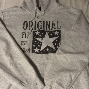 Grå hoodie med tryck och stjärnmotiv - Säljer en grå hoodie med svart tryck från SCANDI  i storlek S framtill där det står 'ORIGINAL FIT EST. 2024' och ett stort stjärnmotiv med mindre stjärnor runtom. Tröjan har huva med dragsko och en klassisk magficka. Perfekt för en avslappnad stil. Tröjan är använd cirka 6 gånger och är i utmärkt skick. Tveka inte att höra av er vid frågor och funderingar. Priset går att diskutera!