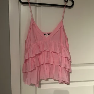Rosa volangtopp med smala axelband - Säljer en söt rosa topp med smala axelband och flera lager volanger. Toppen har v-ringning och är perfekt för varma dagar. Superfin att styla till både jeans och kjol! Aldrig använd så är i bra skick. 
