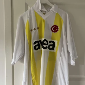 Fenerbahce fotbollströja Alex 20 - Säljer en vit och gul Fenerbahce fotbollströja med tre stjärnor, tryck av 'Alex 20' på ryggen och klubbmärke med turkisk flagga på bröstet. Kortärmad modell med klassisk krage och tryckt sponsorlogga framtill.