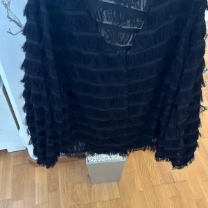 Svart skjorta med fransar - Säljer en svart skjorta med långa ärmar och fransdetaljer över hela plagget. Skjortan har knäppning framtill och en cool, unik look som sticker ut. Perfekt för dig som vill ha något annorlunda i garderoben!