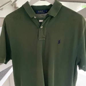 Grön pikétröja från Polo Ralph Lauren - Klassisk grön pikétröja från Polo Ralph Lauren med broderad logga i blått på bröstet. Tröjan har korta ärmar, krage och knäppning med två knappar framtill. Perfekt för en stilren och avslappnad look. Den är i storleken M och är slimfit . Priset är ej hugget i sten!