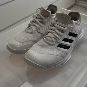 Vita Adidas sneakers med svarta ränder - Säljer ett par fräscha vita Adidas sneakers med de klassiska tre svarta ränderna på sidan. Skorna har snörning och en platt sula, perfekt för en sportig och stilren look.