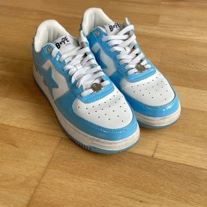 BAPE STA sneakers i blått och vitt - Säljer ett par BAPE STA sneakers i blankt blått och vitt skinn. Skorna har klassisk låg modell med vit och blå sula, rund tå och snörning. BAPE-logga syns på hälen och sidan. Perfekta för dig som gillar streetwear och vill sticka ut med färg.