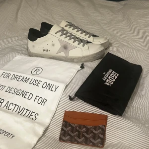 Golden Goose superstar med korthållare  - Säljer ett par vita sneakers från Golden Goose med grå stjärna på sidan och svarta detaljer vid hälen. Skorna har snörning och en platt sula. Perfekta för dig som gillar en stilren och trendig look, funkar speciellt bra nu till sommaren. Storlek 44 men kan även passa 43 eller 45. Korthållaren medkommer vid köp. OBS; lnt 0rglna1 
