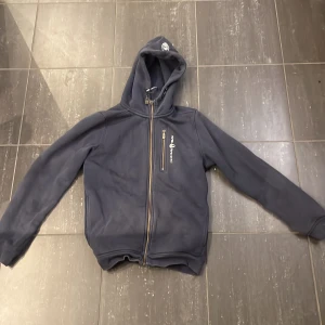 Blå huvjacka från Sail racing - Mörkblå huvjacka från Sail racing med dragkedja och logga på bröstet. Hoodie har huva och långa ärmar, samt en snedställd bröstficka med dragkedja. Perfekt för en sportig och avslappnad stil.