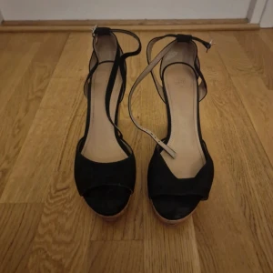 Svarta kilklackar med korksula - Säljer ett par svarta sandaler med kilklack och öppen tå. Skorna har en snygg korksula och remmar runt vristen för extra stöd. Perfekta för sommaren och ger en stilren look till din outfit.