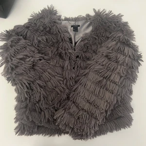 Grå fransad jacka från Lindex - Säljer en unik grå jacka från Lindex med massor av fransar över hela plagget. Jackan har lång ärm och är fodrad på insidan. Perfekt för dig som vill sticka ut och addera något extra till din outfit. Storlek L. I mycket bra skick.