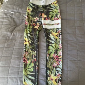 Blommiga träningstights från Stronger - Säljer ett par färgglada träningstights från Stronger i storlek XS. Byxorna har tropiskt blommönster i grönt, gult och rosa samt bred vit midjeresår med logga. Perfekta för gymmet eller löprundan!