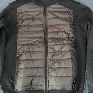 Svart cardigan från Polo Ralph Lauren - Säljer en svart cardigan från Polo Ralph Lauren med quiltad framsida och mjuka ärmar. Jackan har dragkedja, två fickor med dragkedja och röd broderad logga på bröstet. Perfekt för lager-på-lager och stilren look.