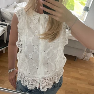 Vit ärmlös blus med spetsdetaljer - Säljer en söt vit ärmlös blus med spets och broderade detaljer. Blusen har krage, knappar framtill och vågig kant nertill. Perfekt till jeans eller kjol för en fräsch look.