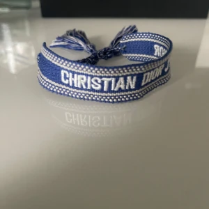Blått armband från Christian Dior - Säljer ett blått armband från Christian Dior med texten 'CHRISTIAN DIOR J'ADIOR' broderad i vitt. Armbandet har knytning och tofsar i ändarna. Perfekt accessoar för att lyfta din stil. Super fet på somrarna och passar all sizer. Skriv till mig vid minsta funderingar!