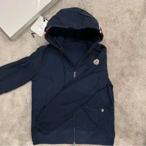  Mörkblå Moncler  vindjacka   - Snygg mörkblå vindjacka från Moncler med huva och dragkedja framtill. Jackan har Moncler-logga på bröstet och en röd, vit och blå rand på huvan. Perfekt för blåsiga dagar och enkel att matcha med det mesta.