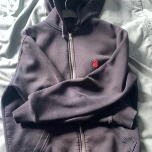 Mörkgrå hoodie med dragkedja från Bloodline - Säljer en svart hoodie från Bloodline i storlek S. Tröjan har dragkedja framtill, huva och en röd broderad detalj på bröstet. Perfekt för en avslappnad stil och enkel att matcha med andra plagg.