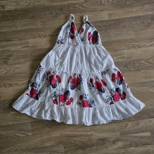 Vit blommig sommarklänning - Söt vit klänning med röda och svarta blommor. Klänningen har smala axelband, v-ringad fram och volangig kjol. Perfekt för varma dagar och har en luftig passform.
