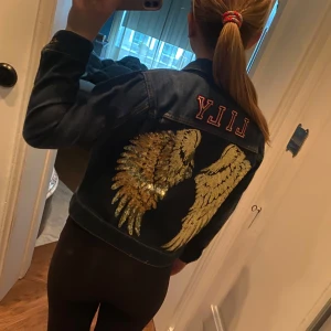 Jeansjacka med guldvingar och namn - Säljer en cool mörkblå jeansjacka med stora glittriga guldvingar på ryggen och namnet 'Lily' i rosa text ovanför. Klassisk modell med knappar framtill och krage. Perfekt statement-plagg för dig som vill sticka ut.